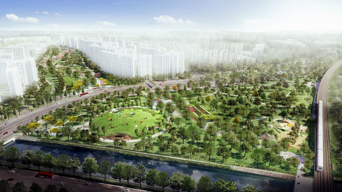 Aerial view of a city park with buildings in the background, surrounded by trees, roads, and a river.